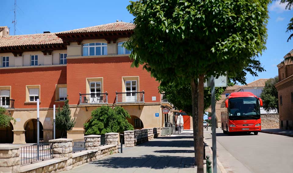 Autobús de excursiones llegando al Hotel Oleum en Belchite, junto a la fachada principal y una calle arbolada del municipio.